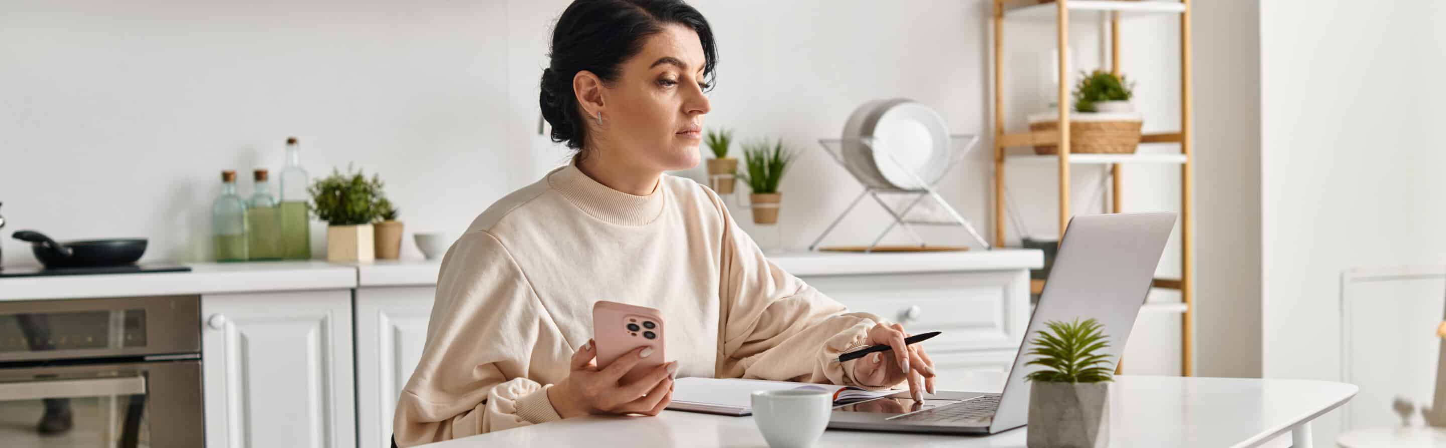 A woman in works from a kitchen table, focused on her laptop and cell phone. A woman working flexible hours at home